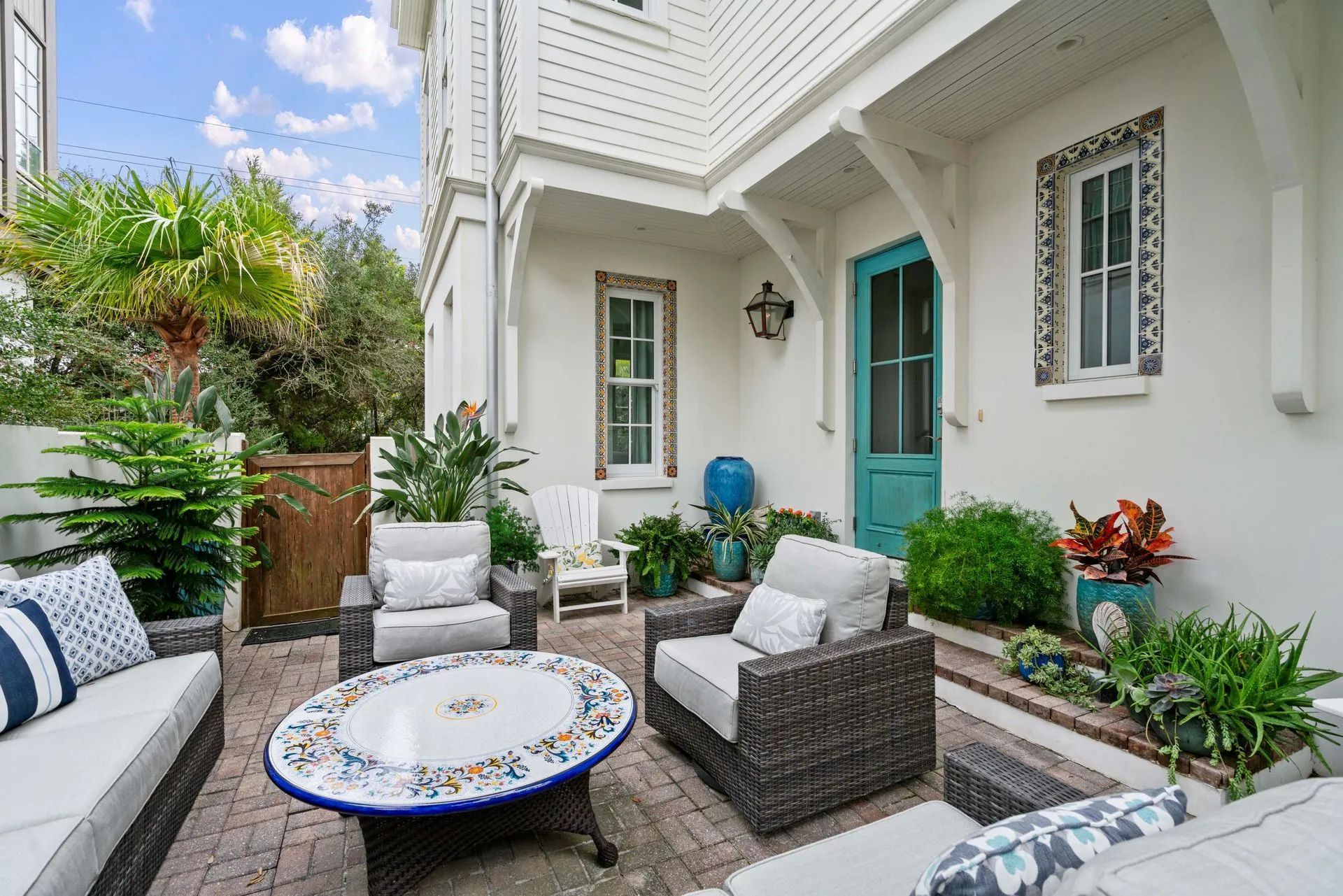 White courtyard patio with wicker seating, round mosaic table, potted plants, and a teal door under a balcony.