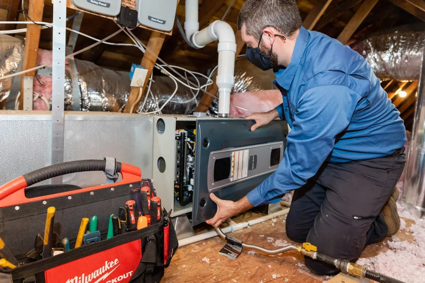 A technician wearing a mask working on a furnace in an attic.