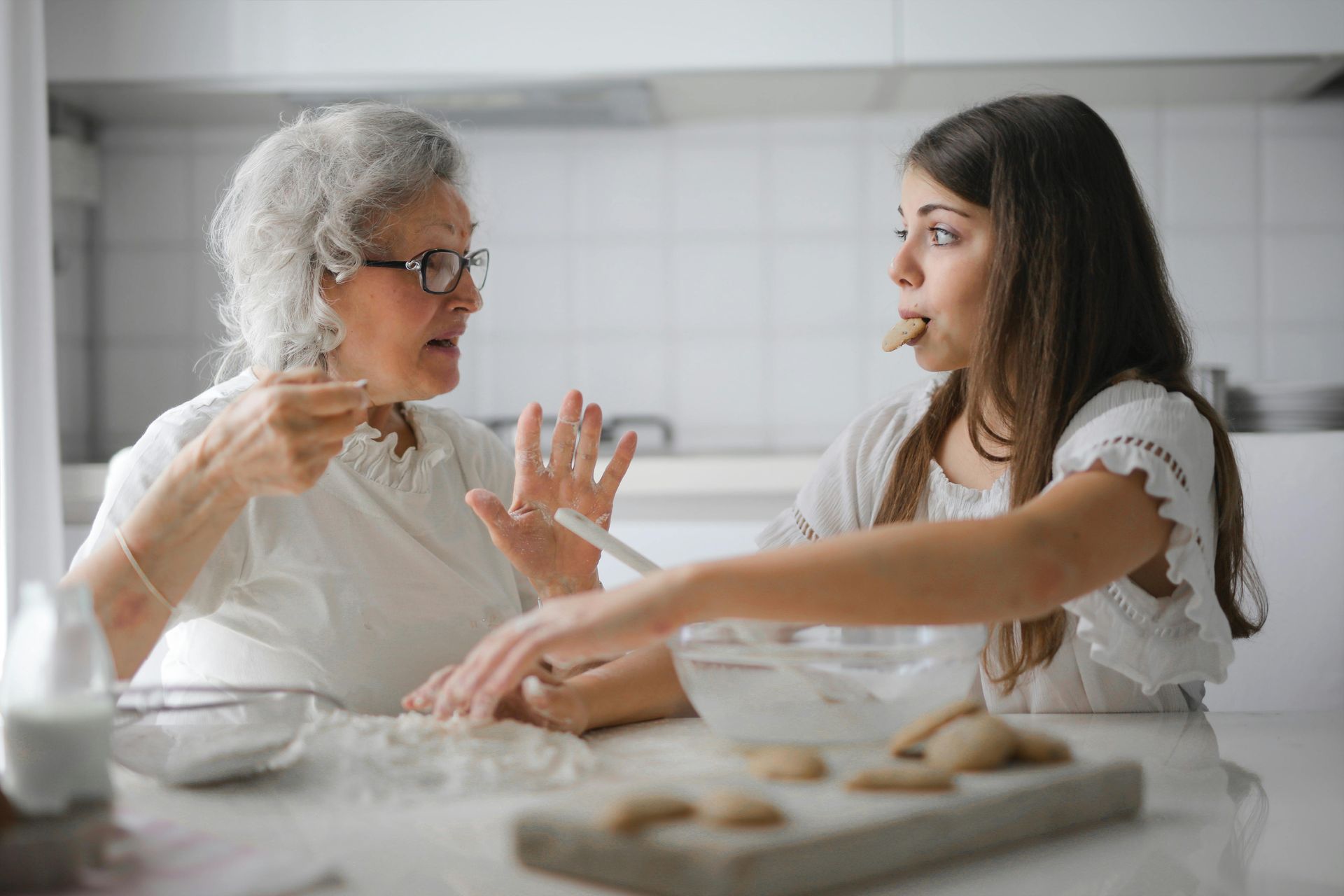 Grandmother and grandchild baking together in a kitchen; the girl tastes batter, the woman gestures.