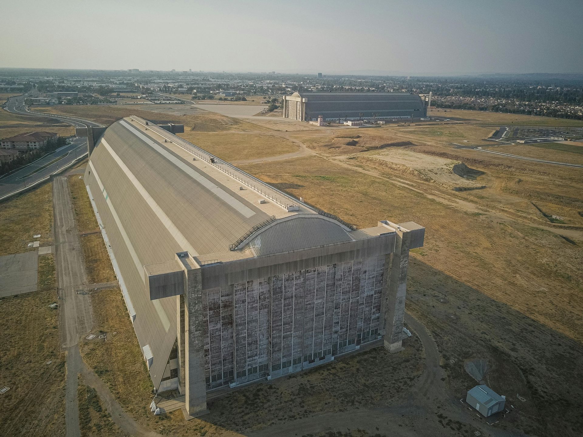 Two large abandoned aircraft hangars on a dry, brown field. Distant town in the background.
