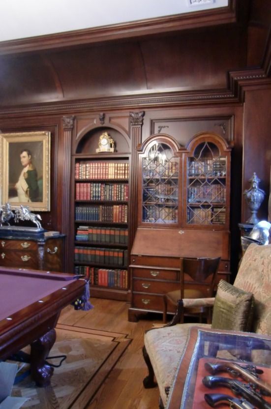 A wood-paneled library with books, a pool table, antique furniture, and a portrait.