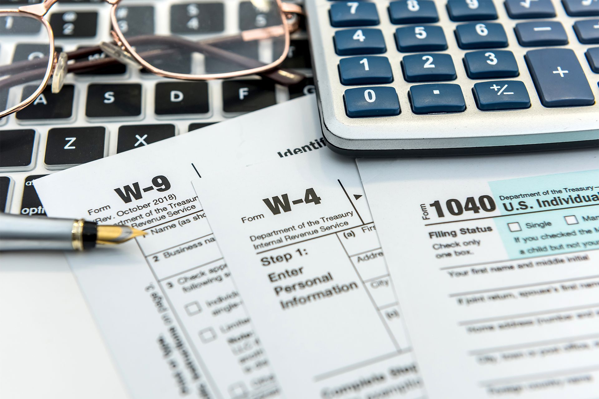 Tax forms W-9, W-4, and 1040 on a laptop with a calculator and glasses, ready for filing.