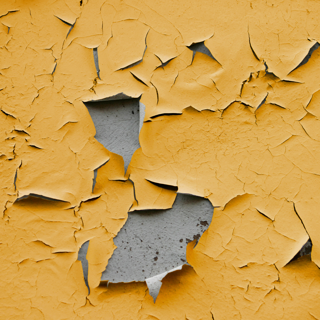 Yellow paint peeling off a gray concrete wall, revealing the surface beneath.