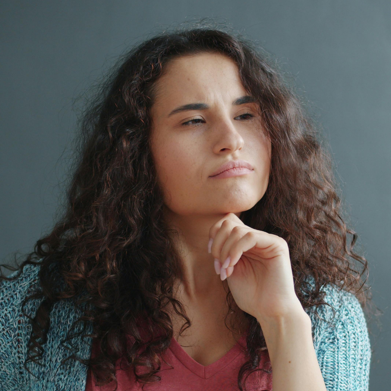 A person with long, curly dark hair against a gray background, resting their chin on their hand with a thoughtful expression.