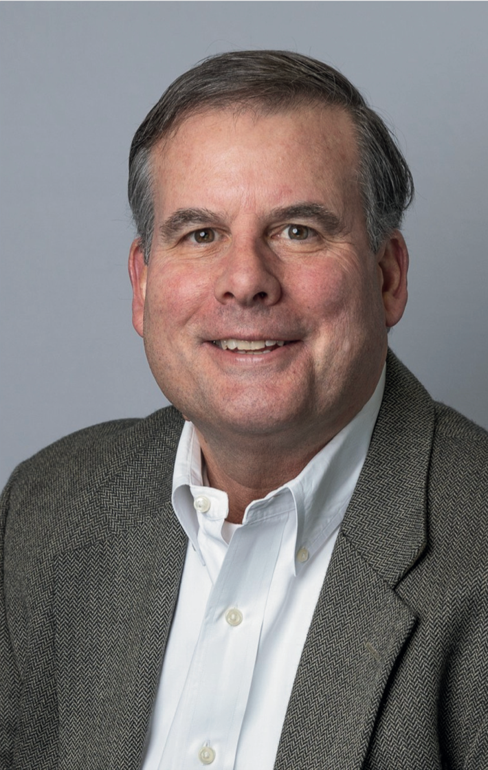 Man in a patterned jacket and open-collared white shirt smiles; studio background.