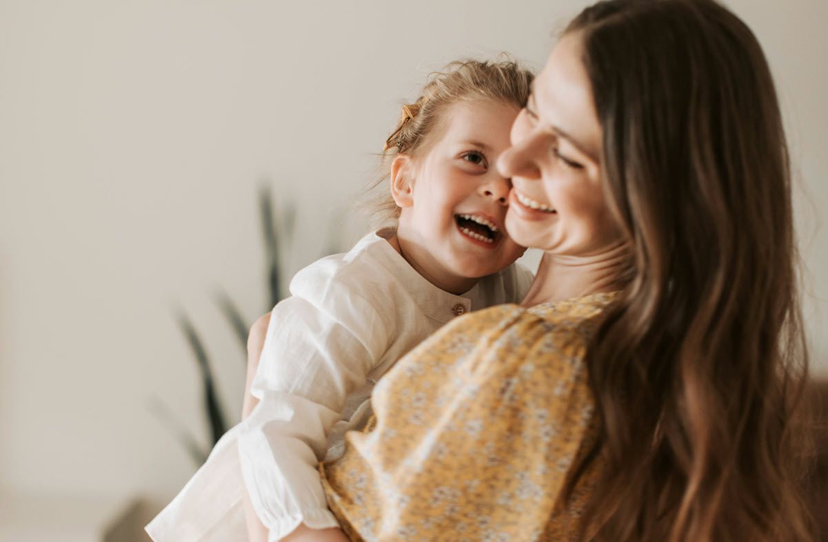 Woman holding and smiling at a laughing child; soft yellow and white clothing, neutral background.