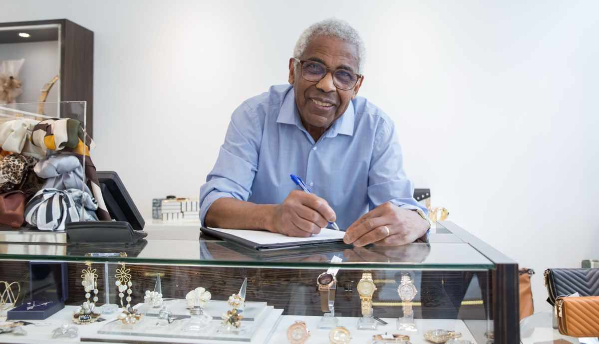Man smiling behind a glass display counter in a bright shop, holding a pen and papers.