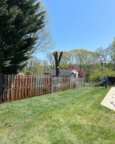 A wooden picket fence borders a lawn, with a topped tree, a small house in the background, and a person standing nearby.