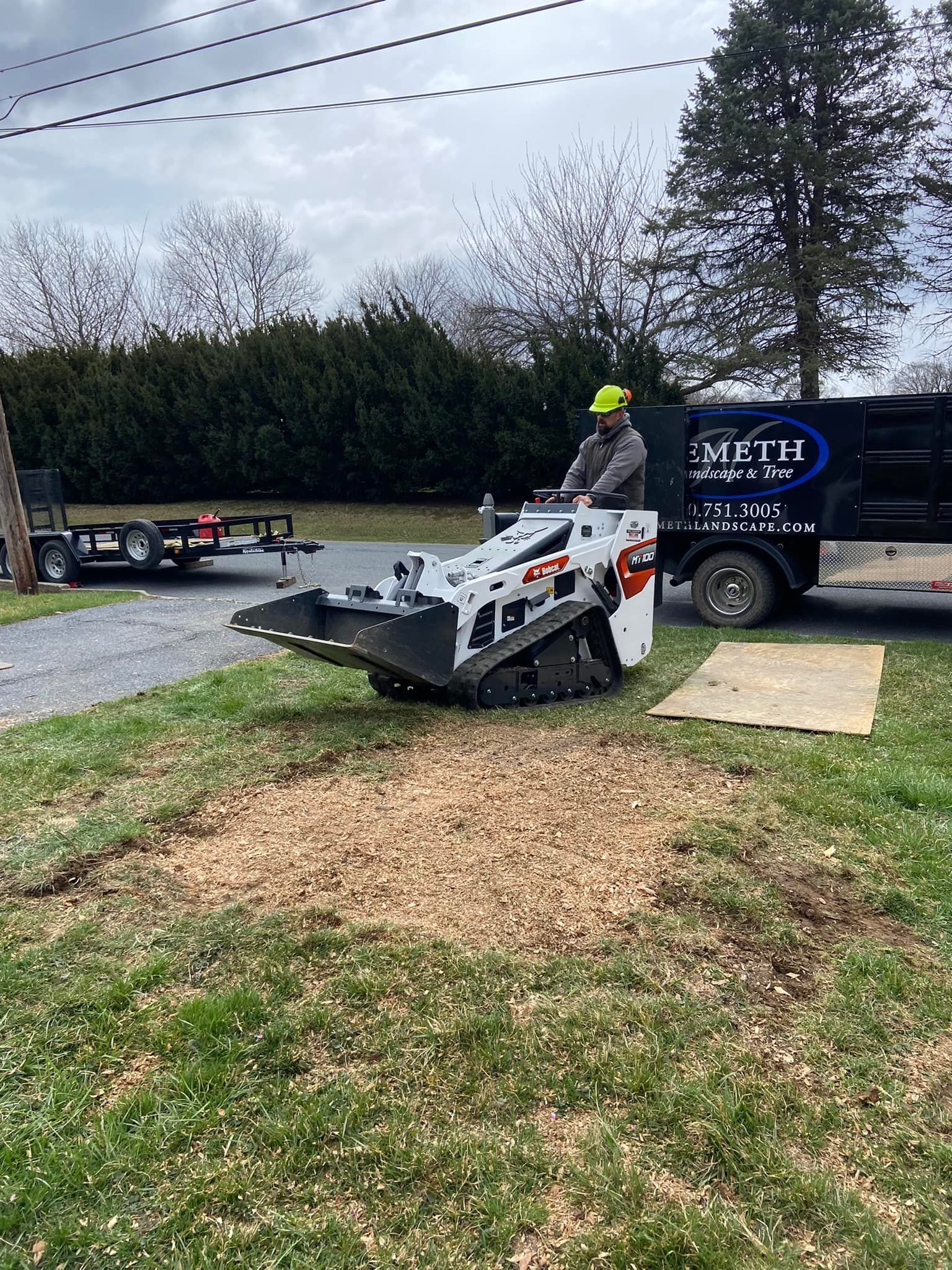 A worker operates a white Bobcat stump grinder on a residential lawn next to a utility trailer.