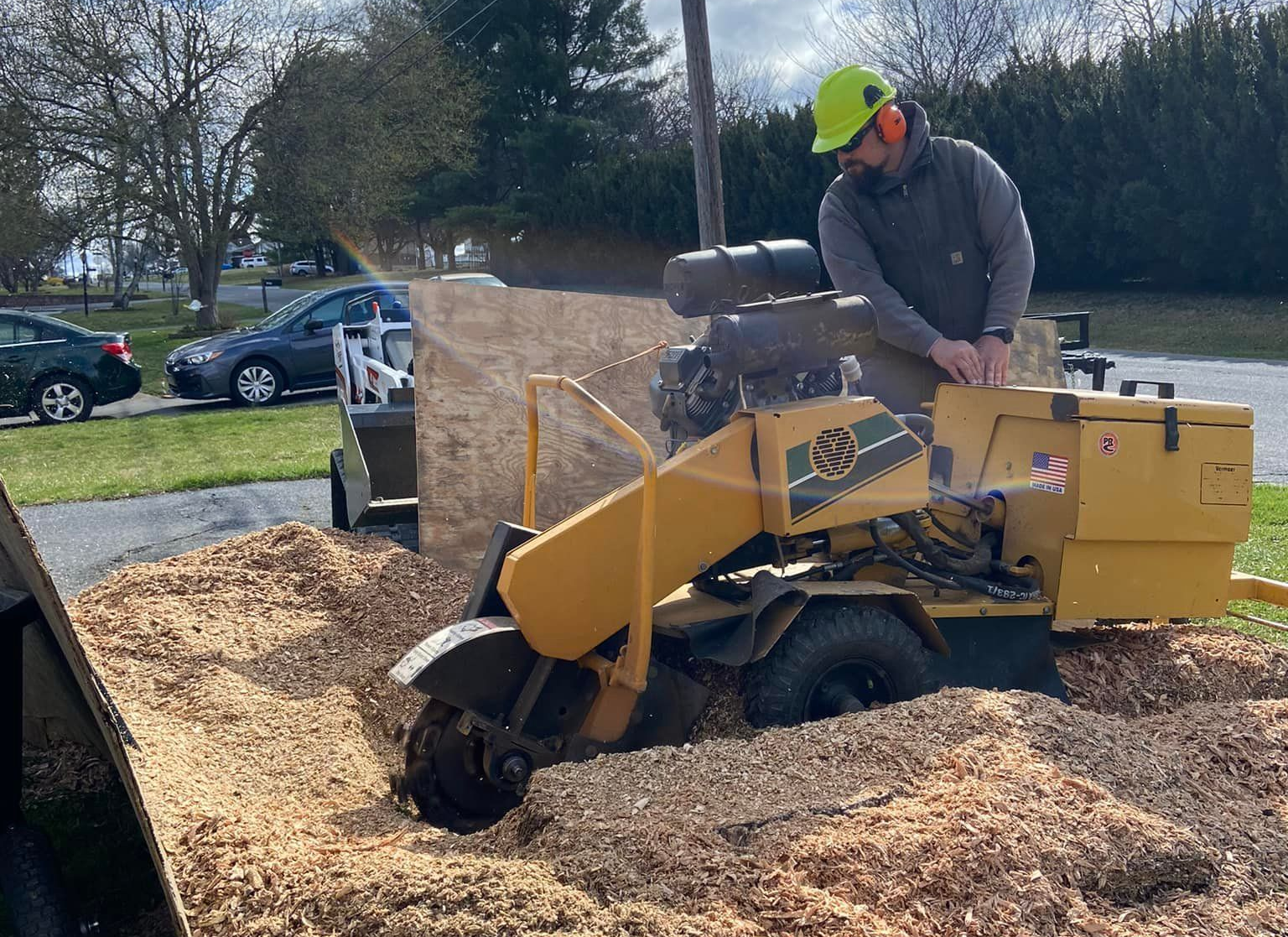 A worker in a high-visibility helmet operates a yellow stump grinder on a pile of wood chips in a residential area.