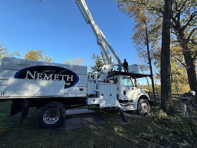 A white Nemeth utility truck with a raised boom bucket in a yard, with a worker in the bucket and another on the ground.