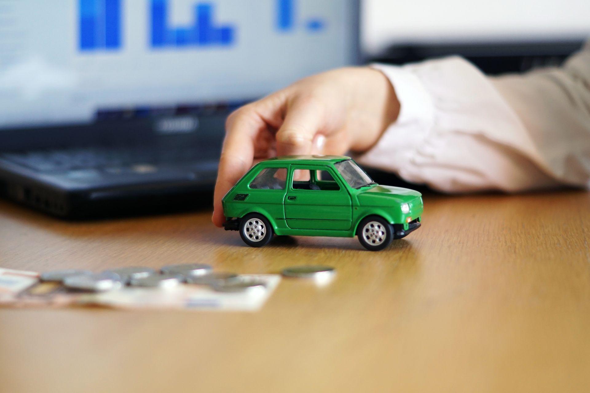 Hand reaching for a green toy car on a desk, with money and a laptop computer.