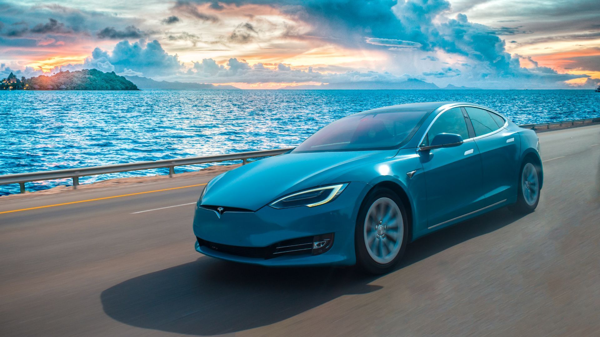 a blue Tesla model S electric vehicle is driving down a road next to the ocean.