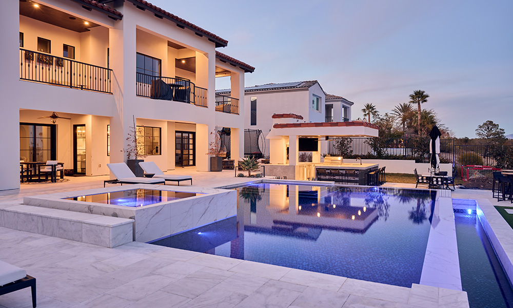 Luxury villa with lit pool at dusk, reflecting modern white buildings and warm patio lights