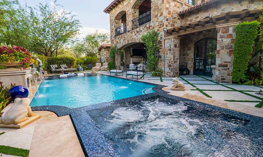 Stone house with arched patio beside a blue backyard pool and landscaped garden