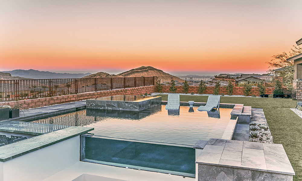 Modern patio with a reflective infinity pool at sunset, overlooking distant hills and a wooden pergola.