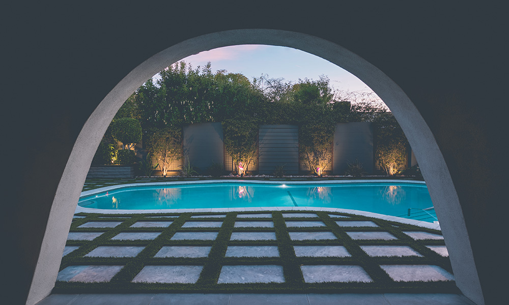 Curved patio opening onto a lit pool at dusk, with arched hedges and a tiled walkway in the foreground