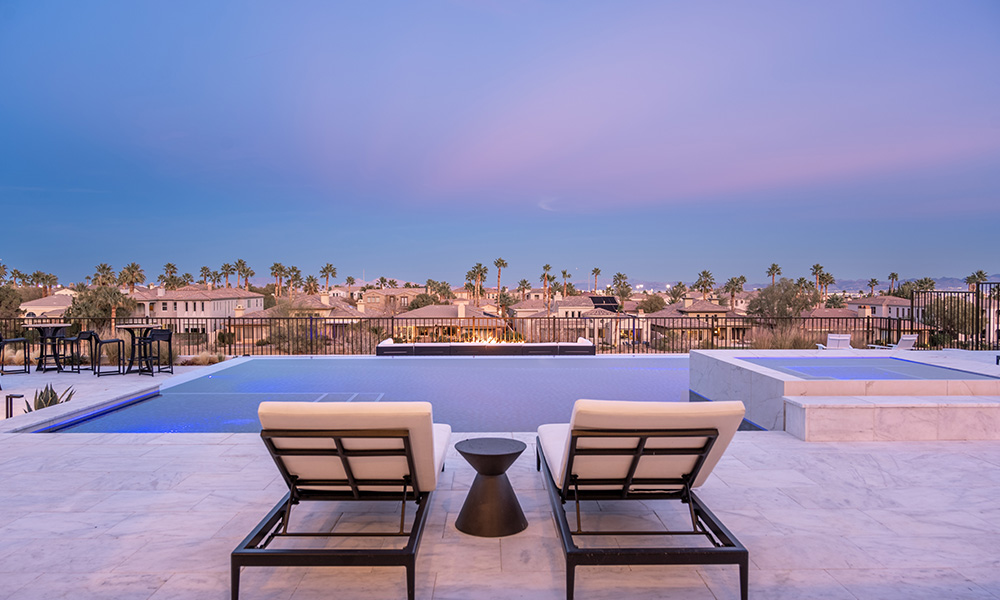 Two lounge chairs by a pool overlooking palm trees at sunset, with a pastel sky.