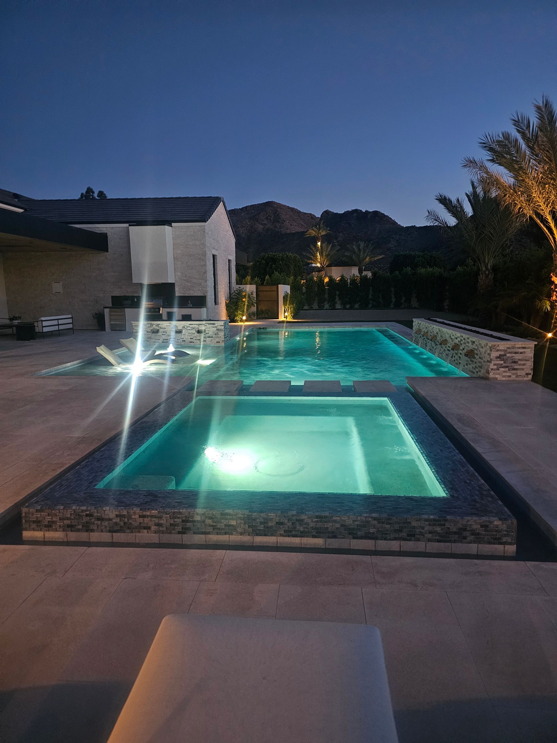 Lit backyard pool and hot tub at dusk beside a modern house with mountains in the background