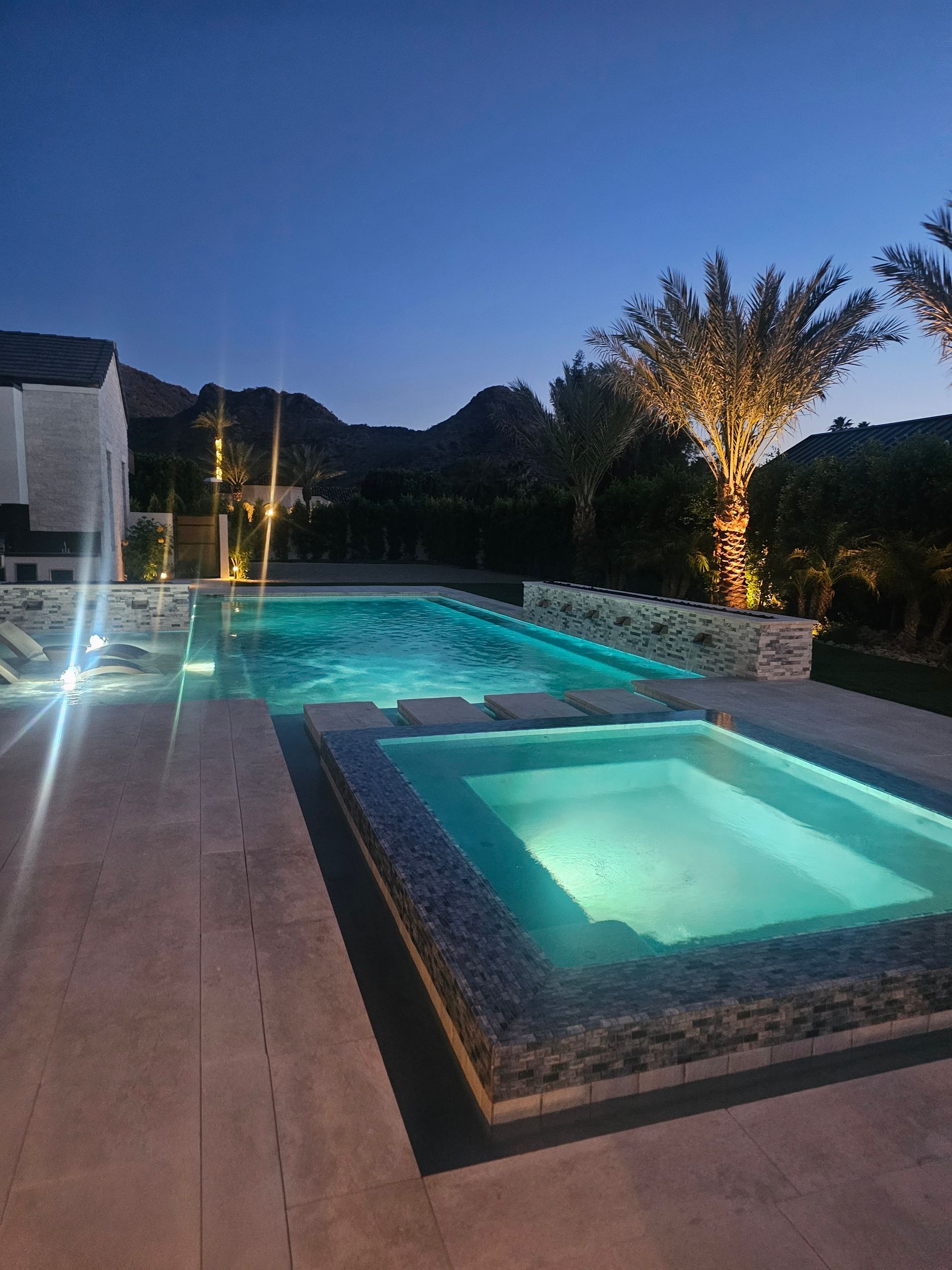 Lit pool and spa beside a deck at dusk, with a glowing tree and dark hills in the background