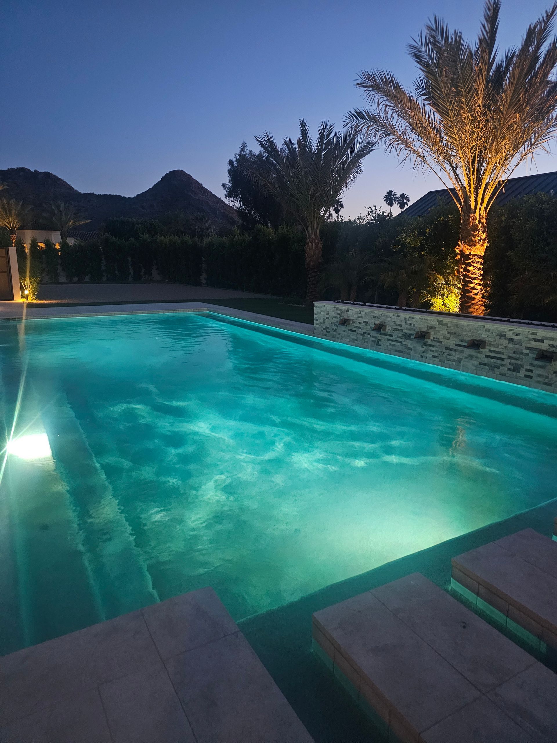 Illuminated backyard pool at dusk with turquoise water and palm trees against a mountain backdrop