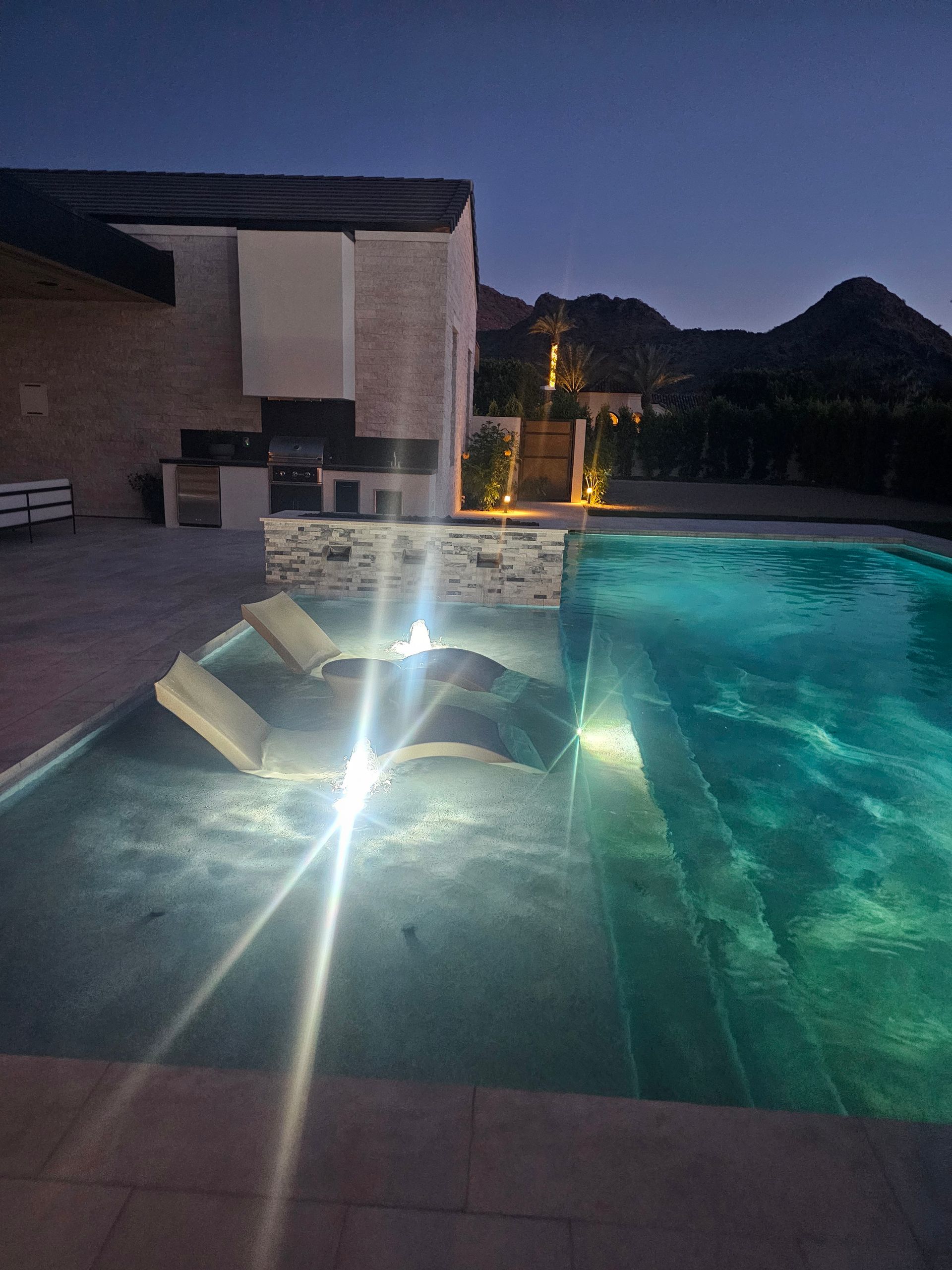 Pool with built-in spa at dusk, turquoise water glowing beside patio seating and desert mountains.