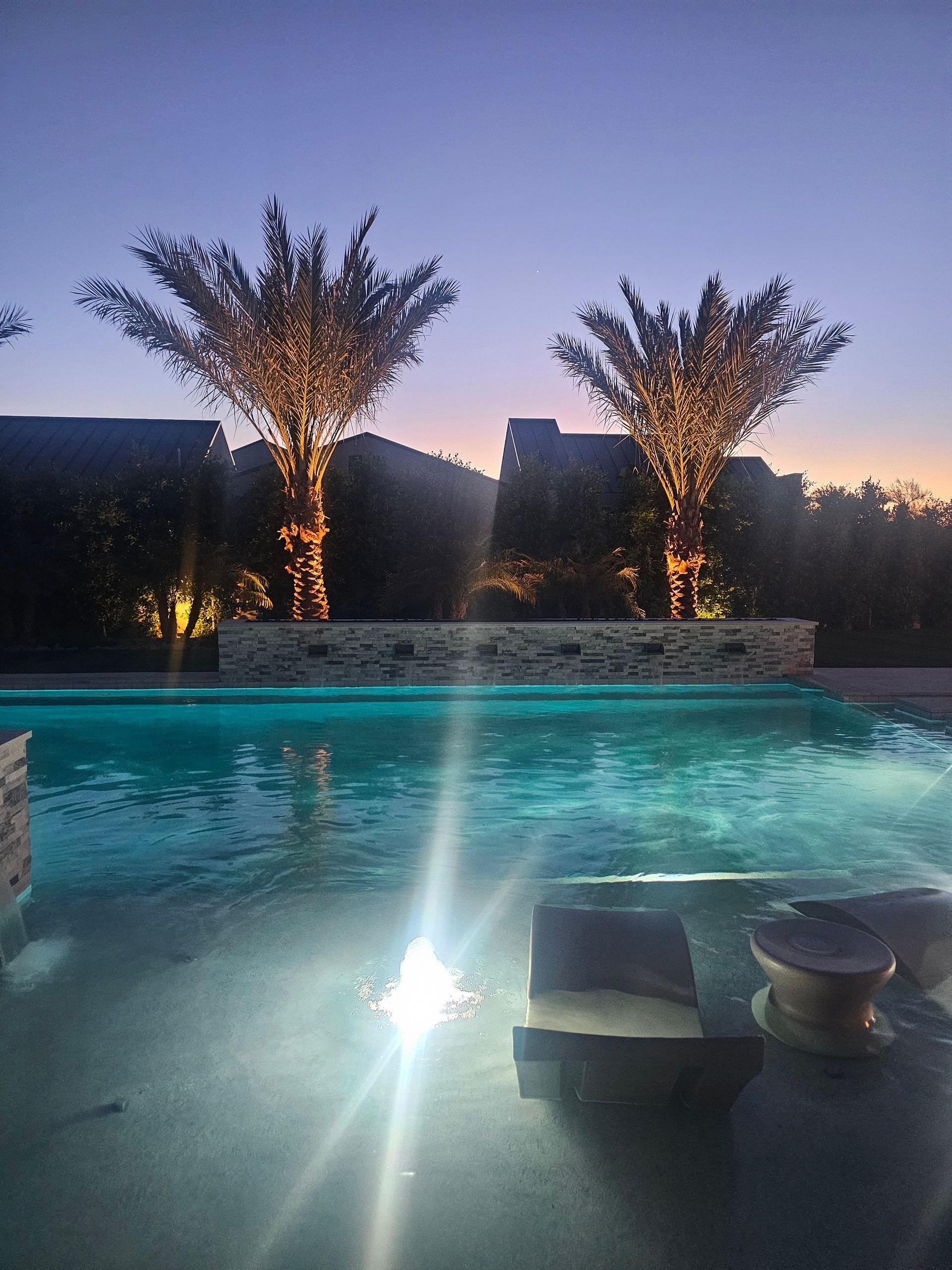 Pool at dusk with illuminated water jets and lit trees beside a stone deck.