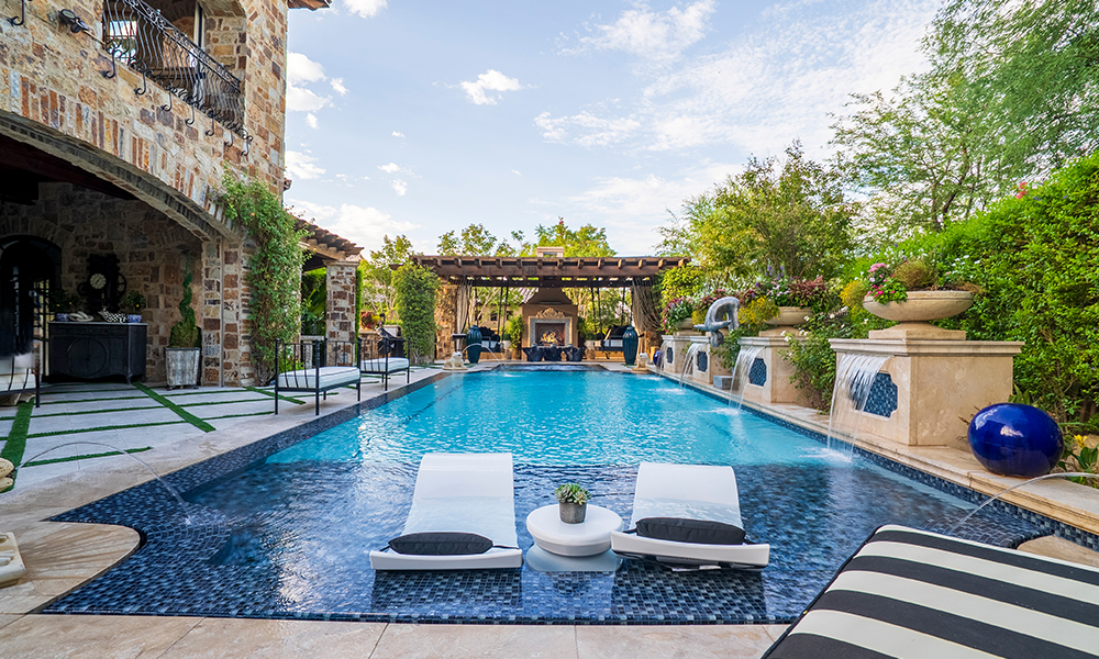 Luxury backyard pool with blue water, striped loungers, and stone patio under a partly cloudy sky