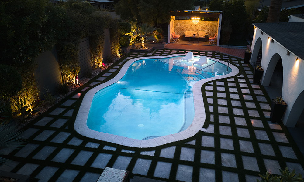 Nighttime backyard pool with blue water, lit by warm lights, beside a tiled patio and white arches.