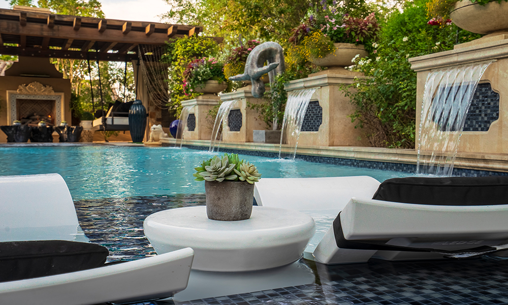 Luxury poolside lounge with white chairs, a potted plant, and cascading wall fountains under a pergola