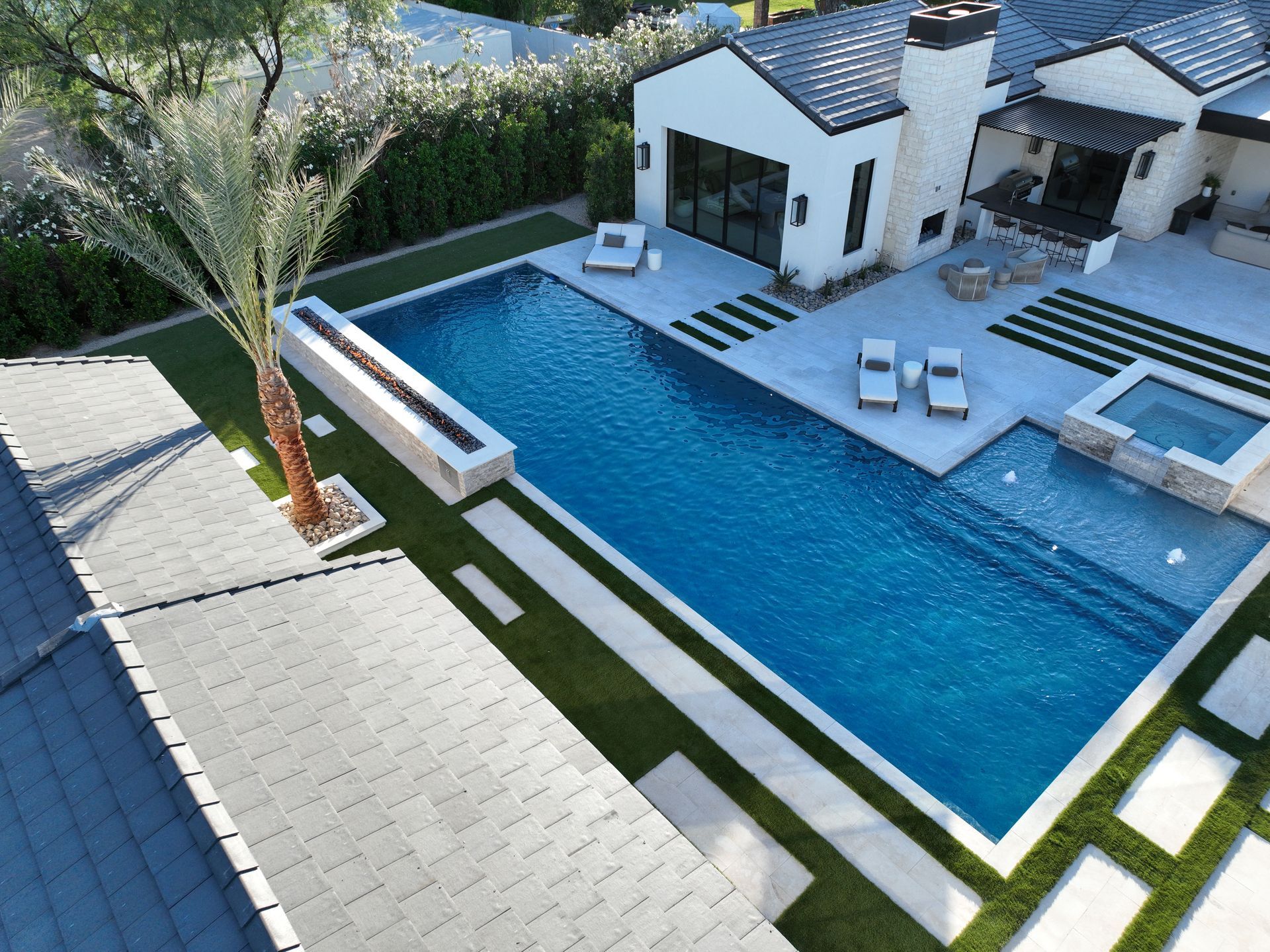 Modern backyard with a blue lap pool, white patio seating, and a small house beside palm trees.
