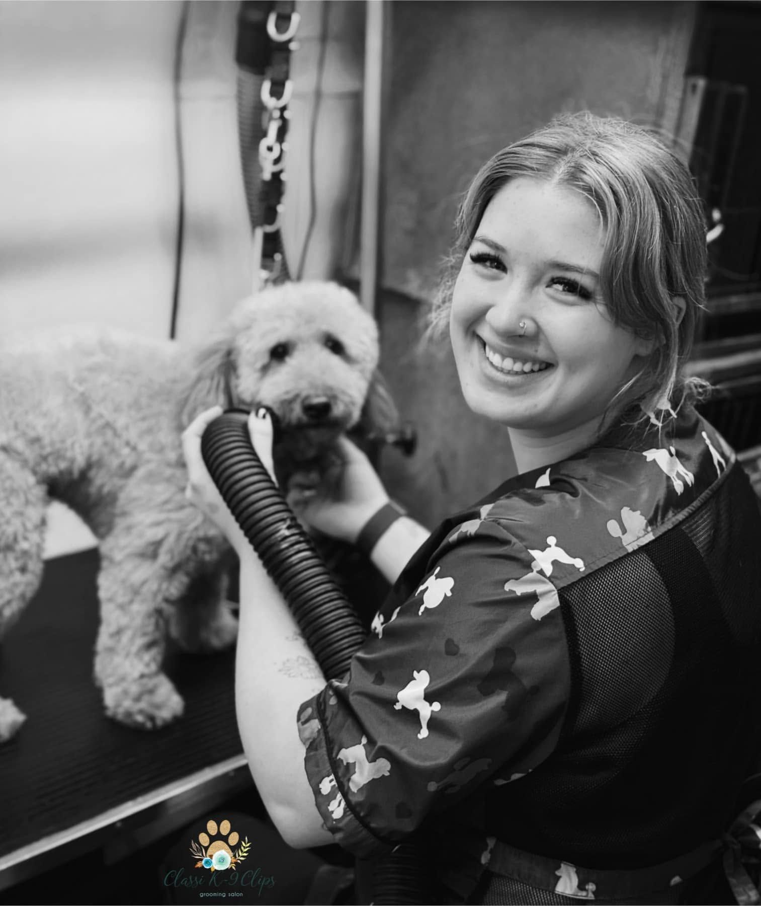 Smiling groomer holding a poodle at a grooming table, with a vacuum hose nearby