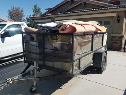 Trailer loaded with rolled rugs and other items, parked in a driveway next to a white truck.