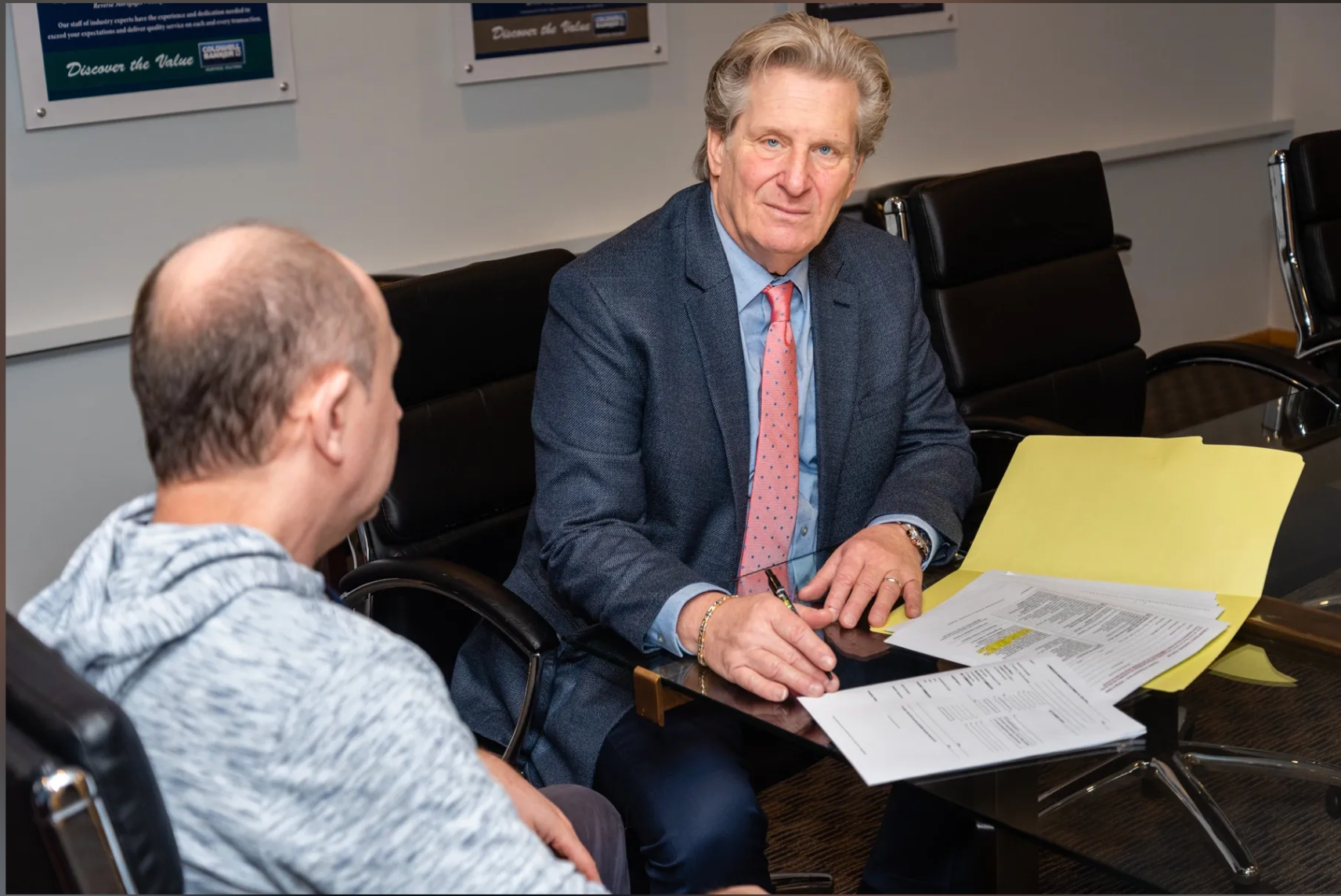 A man in a suit and tie is sitting at a table talking to another man.