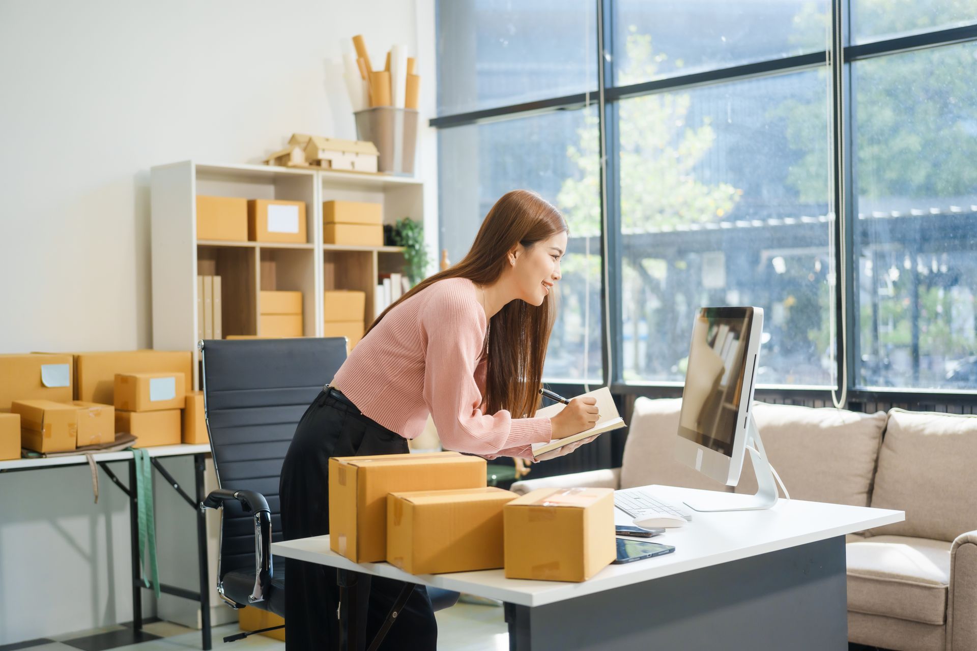 Woman packing boxes at a desk in a bright office, leaning toward a computer monitor.