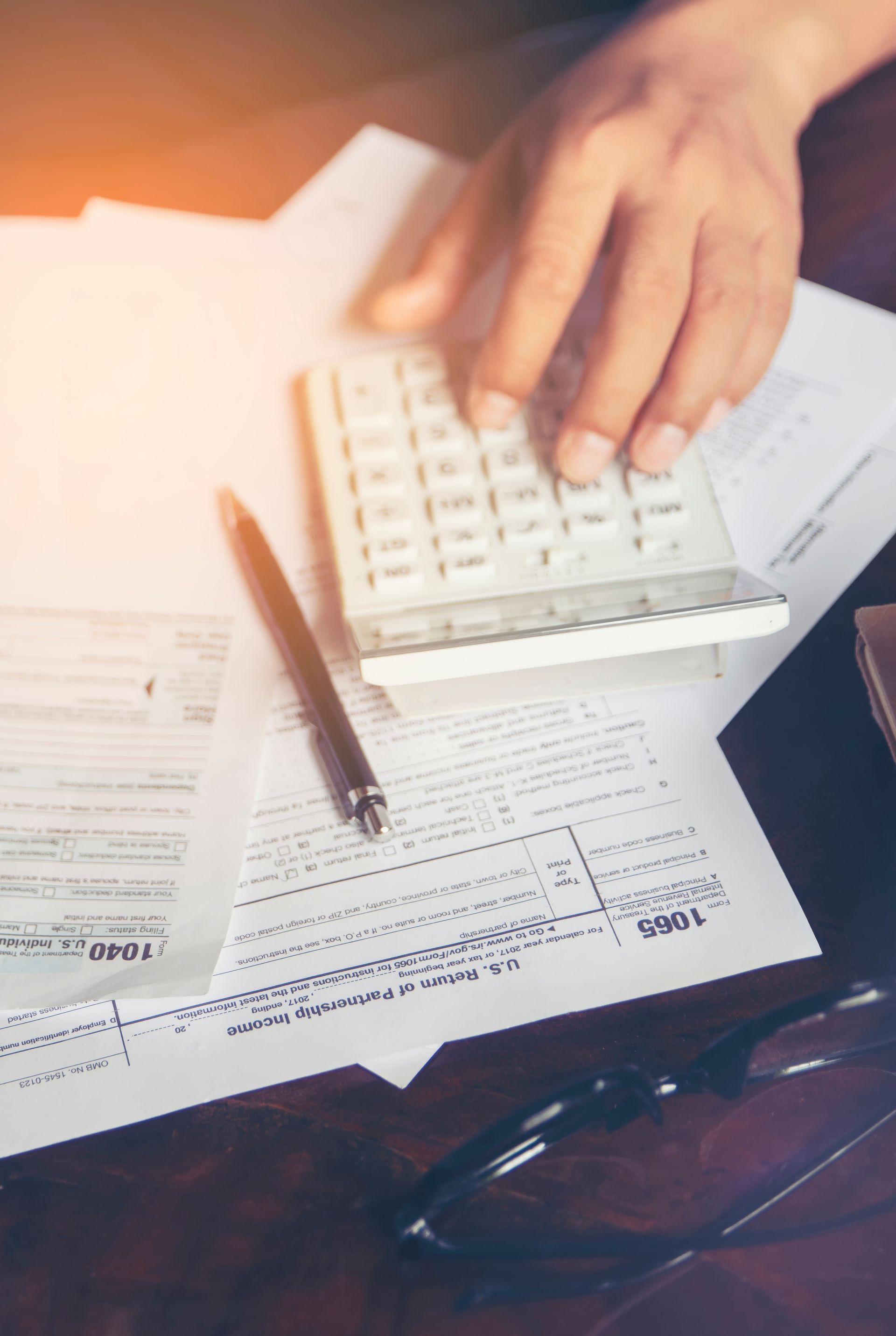 Hand placing calculator on scattered paperwork beside a pen and eyeglasses on a desk