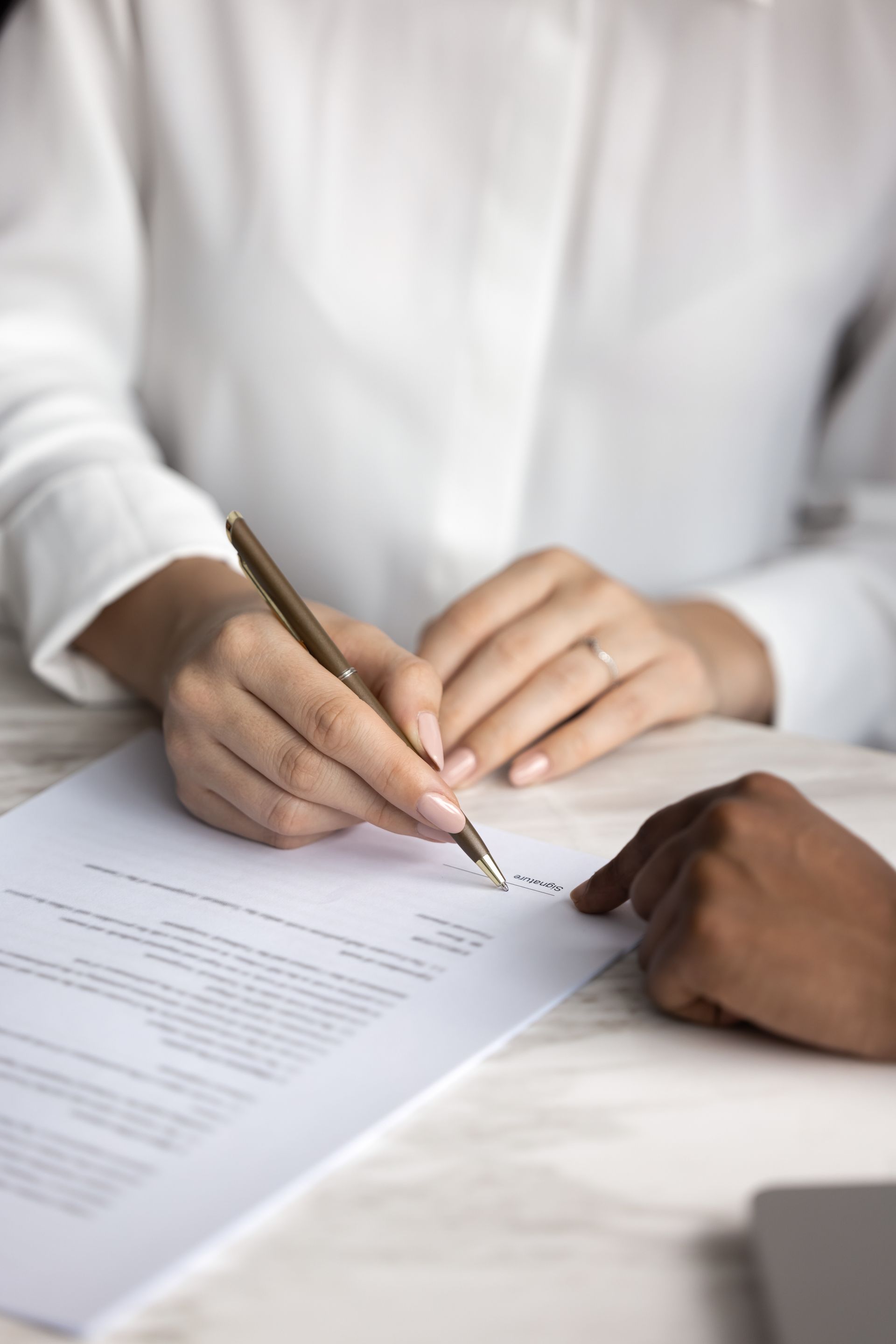 Hands signing a document at a desk, with a third hand pointing at the paper.