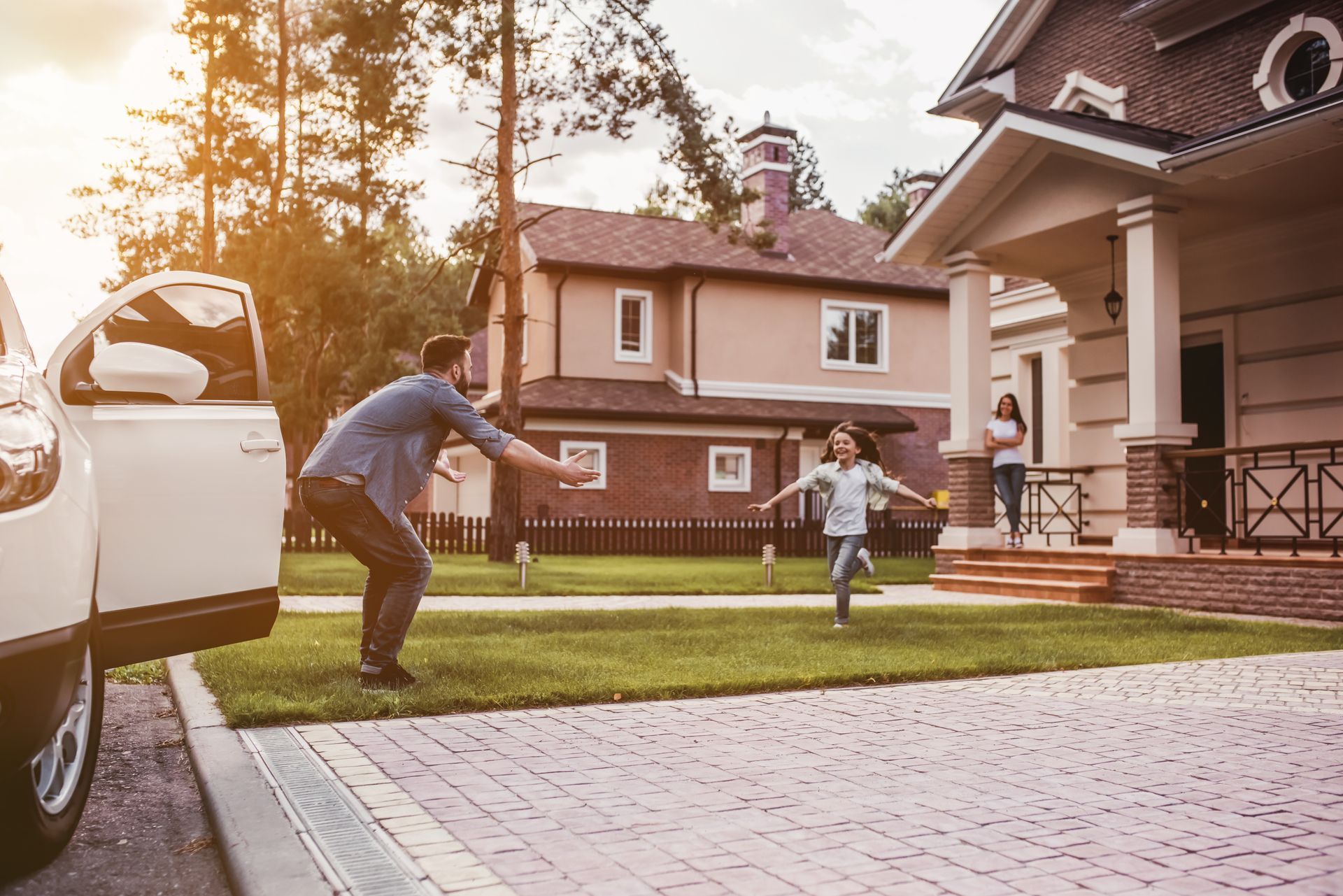 Person tossing a ball to a child on a suburban lawn beside parked car and house at sunset