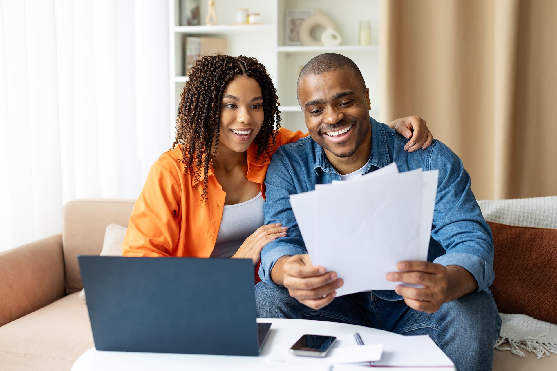 Two people smiling on a couch reviewing papers beside a laptop and phone in a bright living room