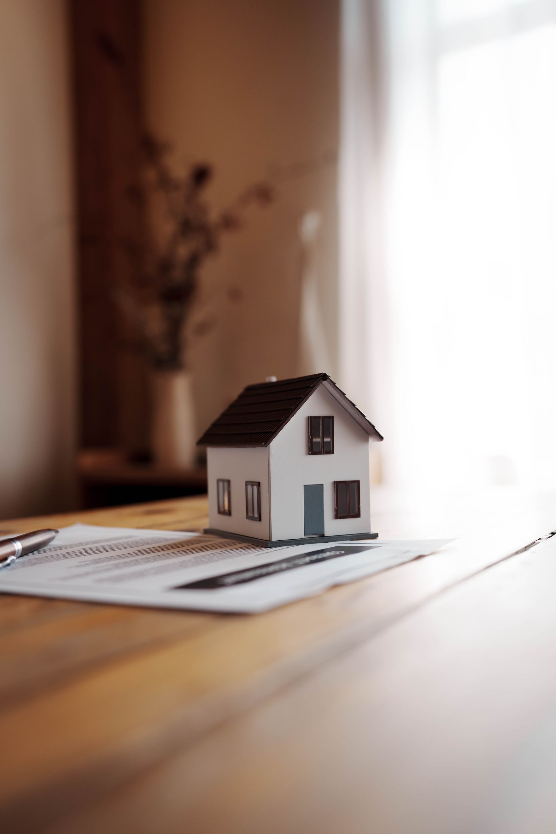 Small white model house on a table near a bright window, with a blurred vase in the background