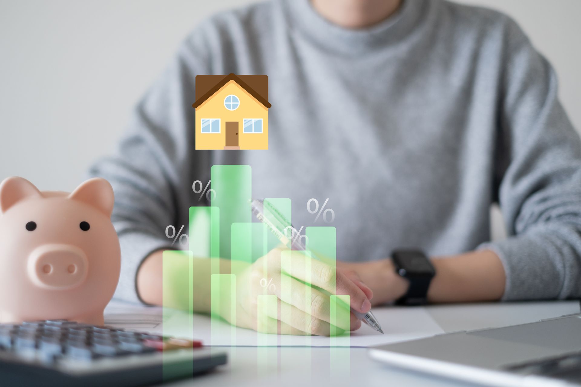 Person at desk with piggy bank, house model, and financial chart overlay on laptop screen