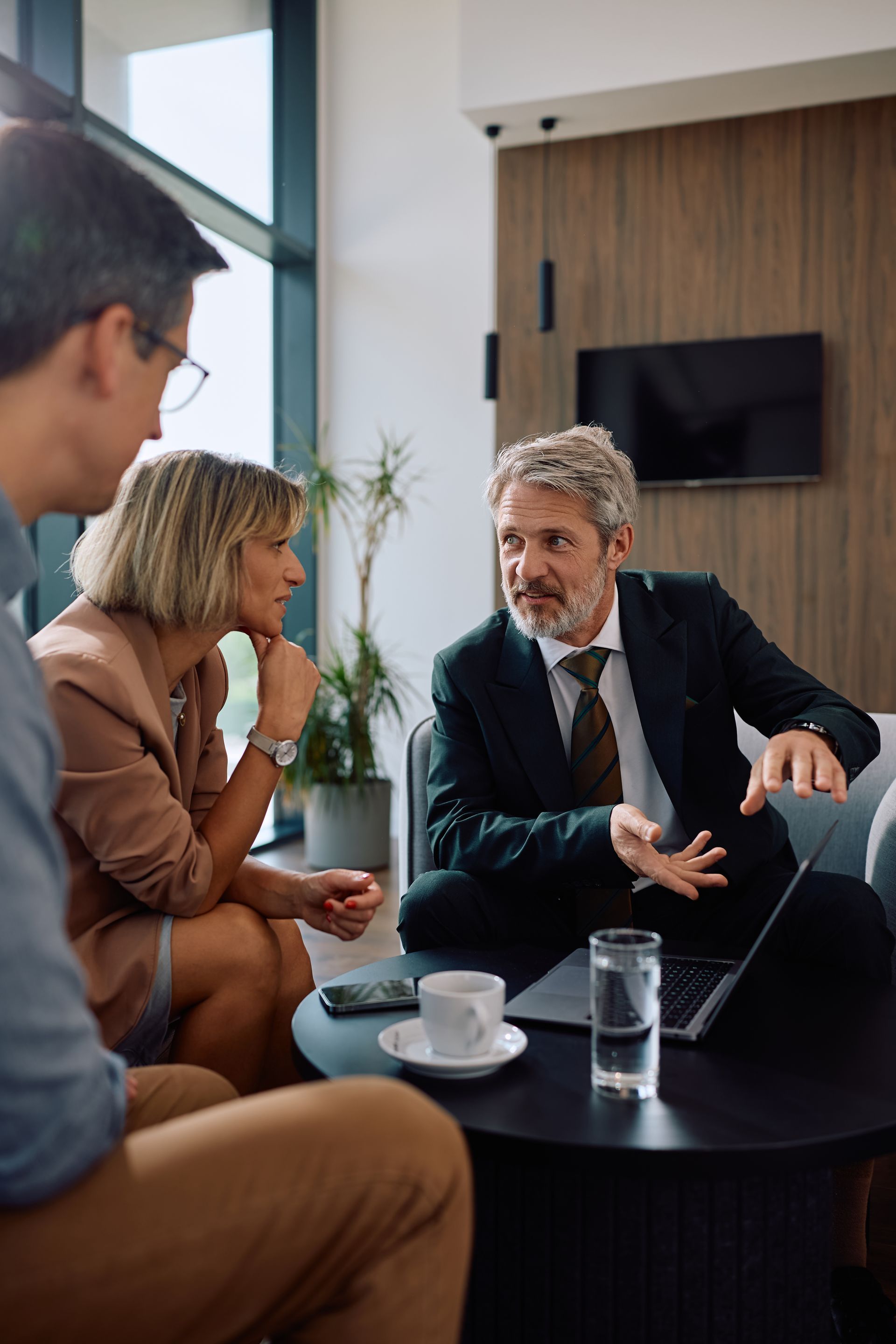 Three people in a modern office lounge discussing over coffee and a laptop