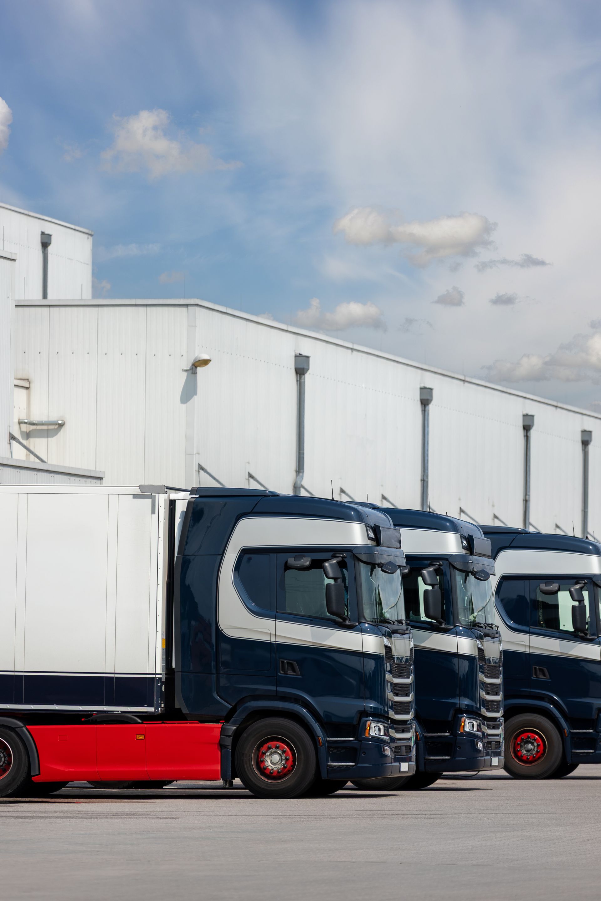 Parked blue and red semi trucks lined up beside a white warehouse under a partly cloudy sky