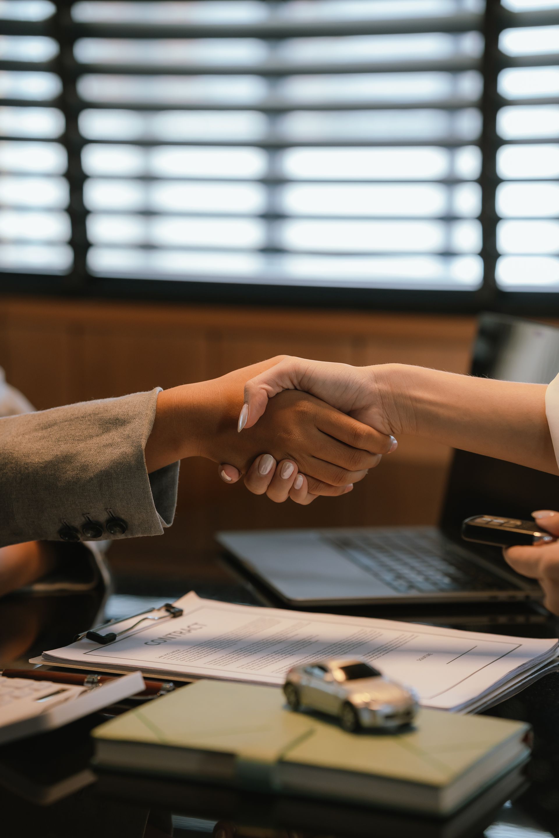 Two people shaking hands across a desk with a notebook, car key, and laptop in a meeting room