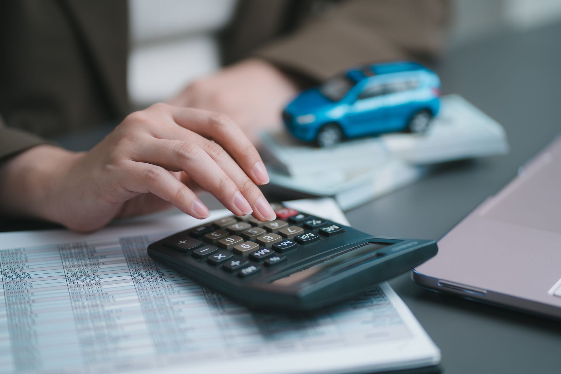 Hand using a calculator beside paperwork, a small blue car model, and a laptop on a desk