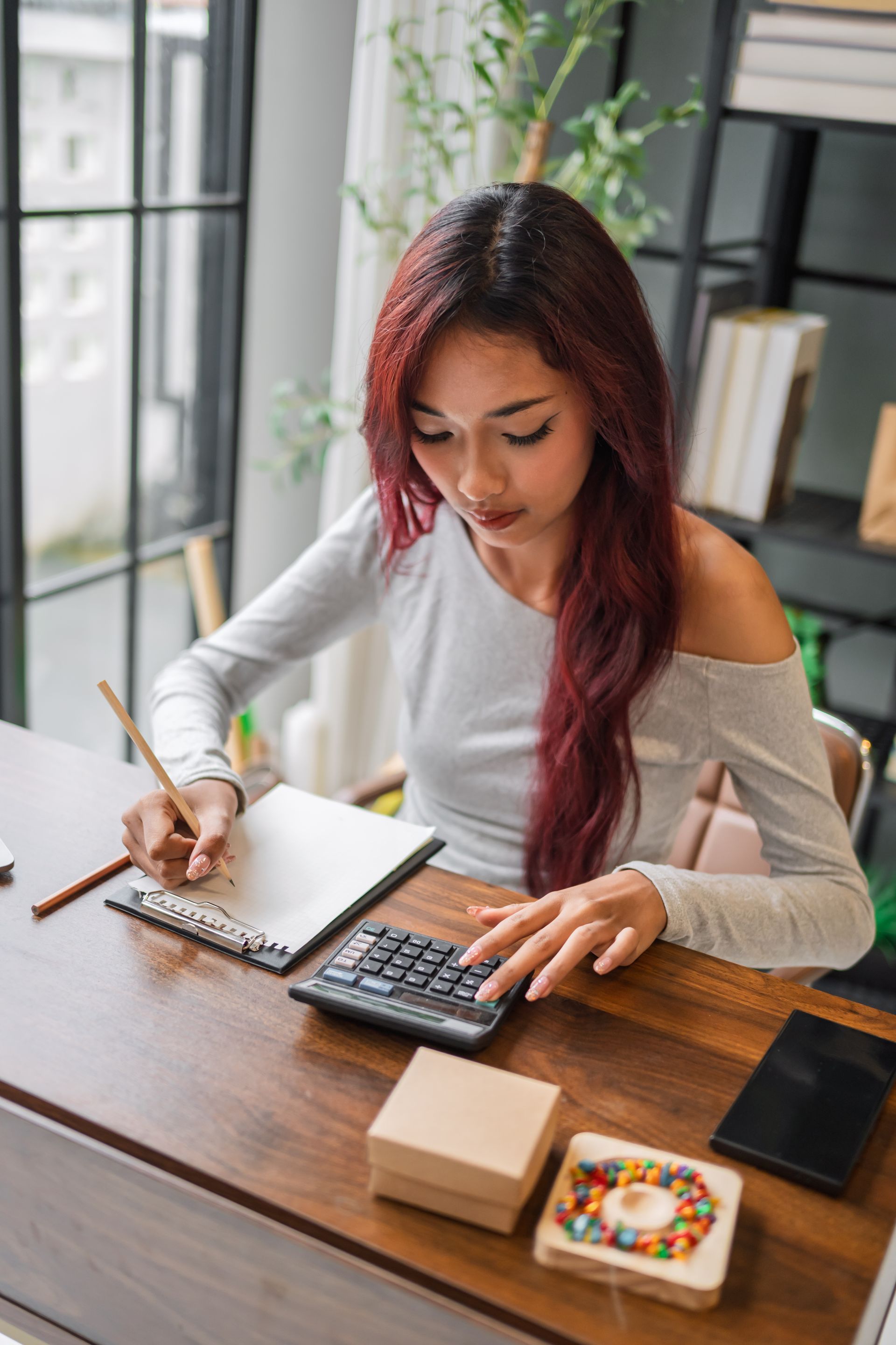 Woman writing in a notebook at a desk with a calculator, laptop, and tablet by a window