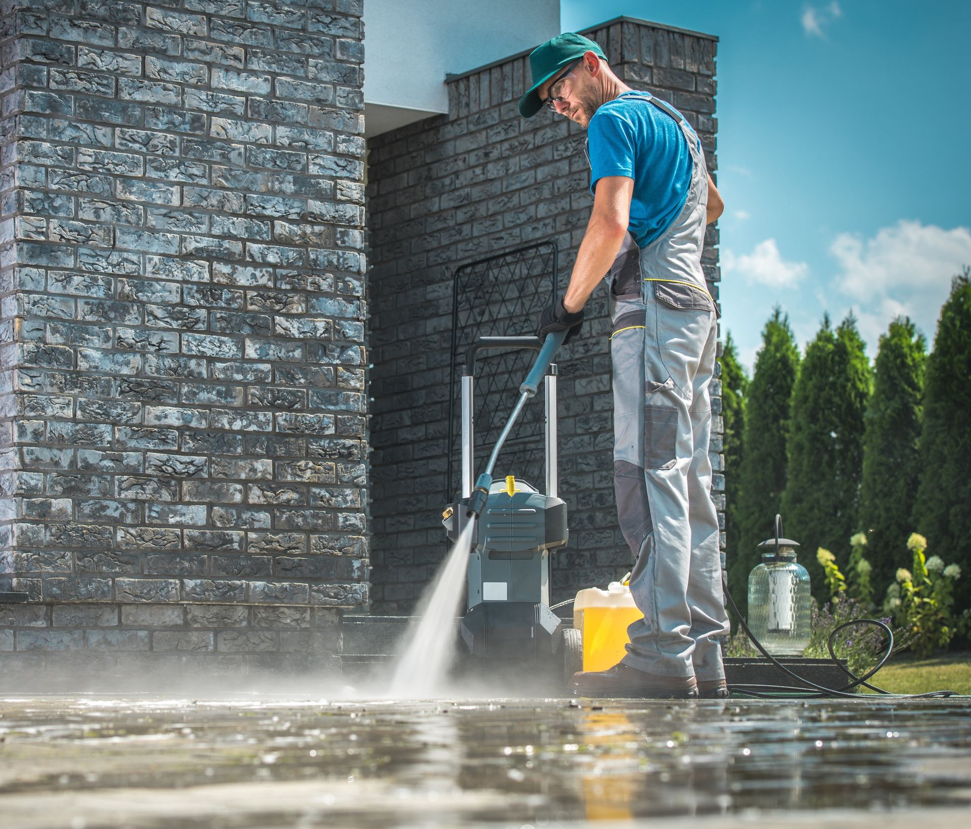 Worker pressure washing a brick patio beside a house, spraying water onto the wet ground.