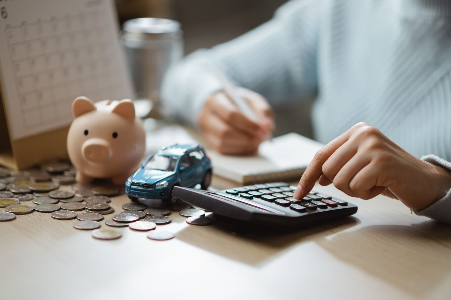 Person using calculator beside coins, toy car, and piggy bank at a desk