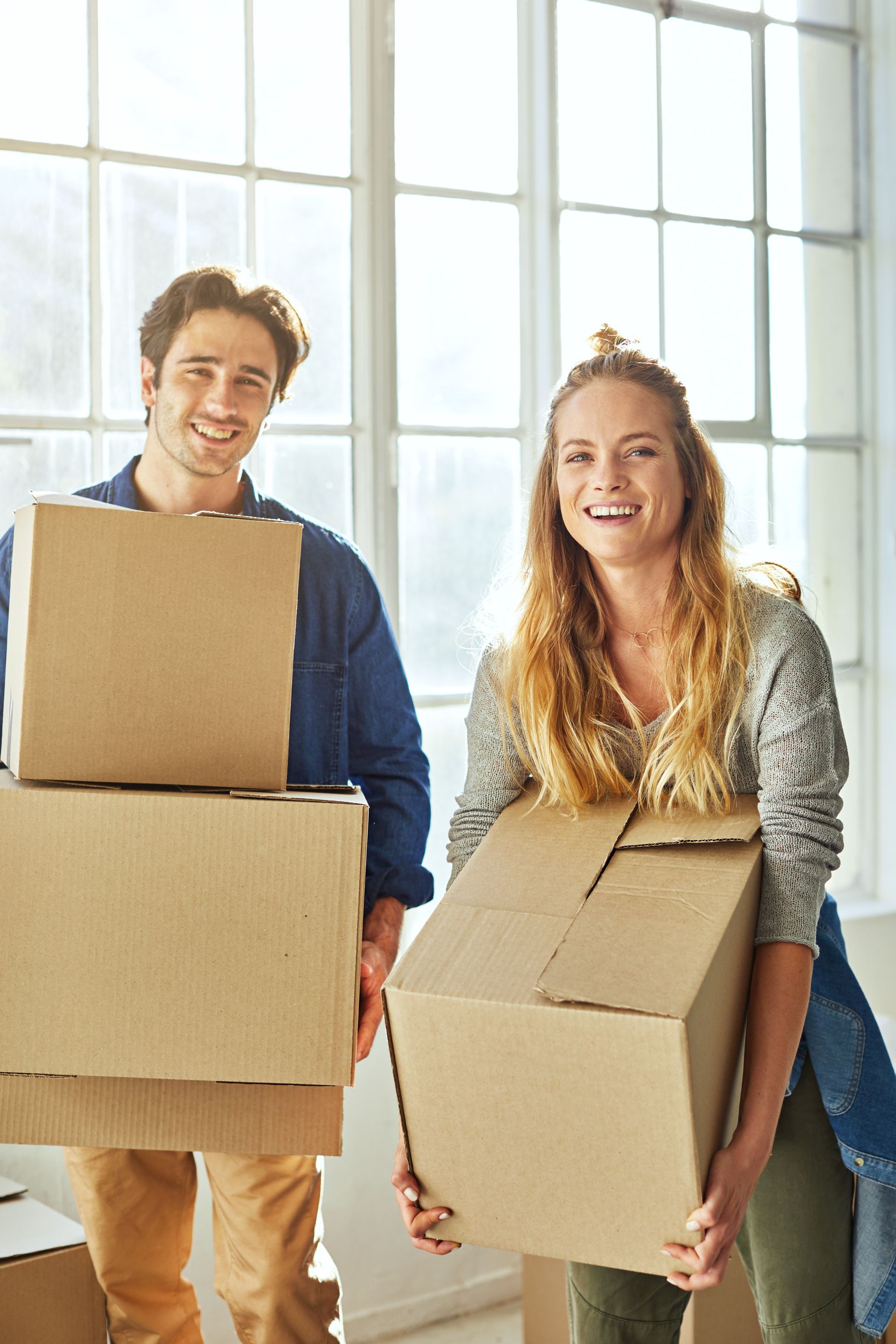 Two people carrying moving boxes in a bright room with large windows