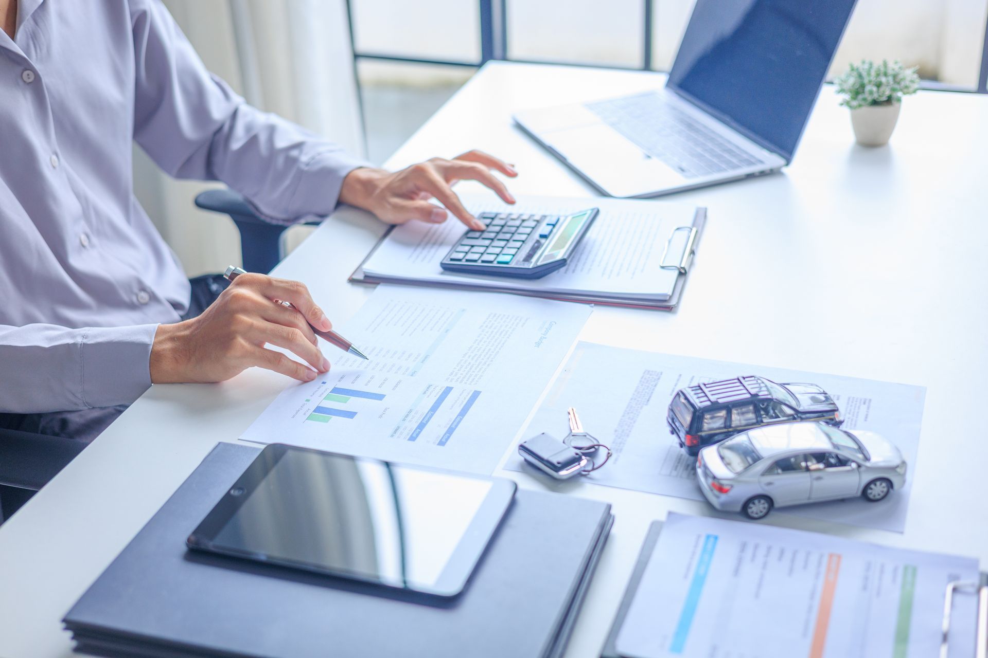 Hands using calculator at a desk with laptop, tablet, documents, and toy car model